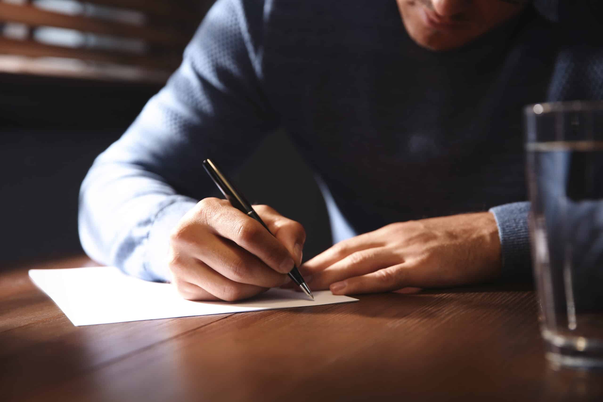 A man in a blue sweater writing on a white sheet of paper at a wooden table, with a glass of water beside him, focusing on rural economies.