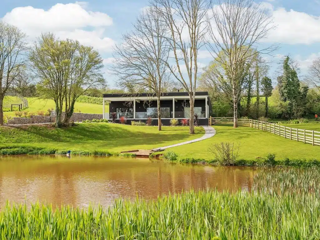 Modern house with large windows overlooking a pond, surrounded by green fields and trees under a cloudy sky. A gravel path leads to the house.