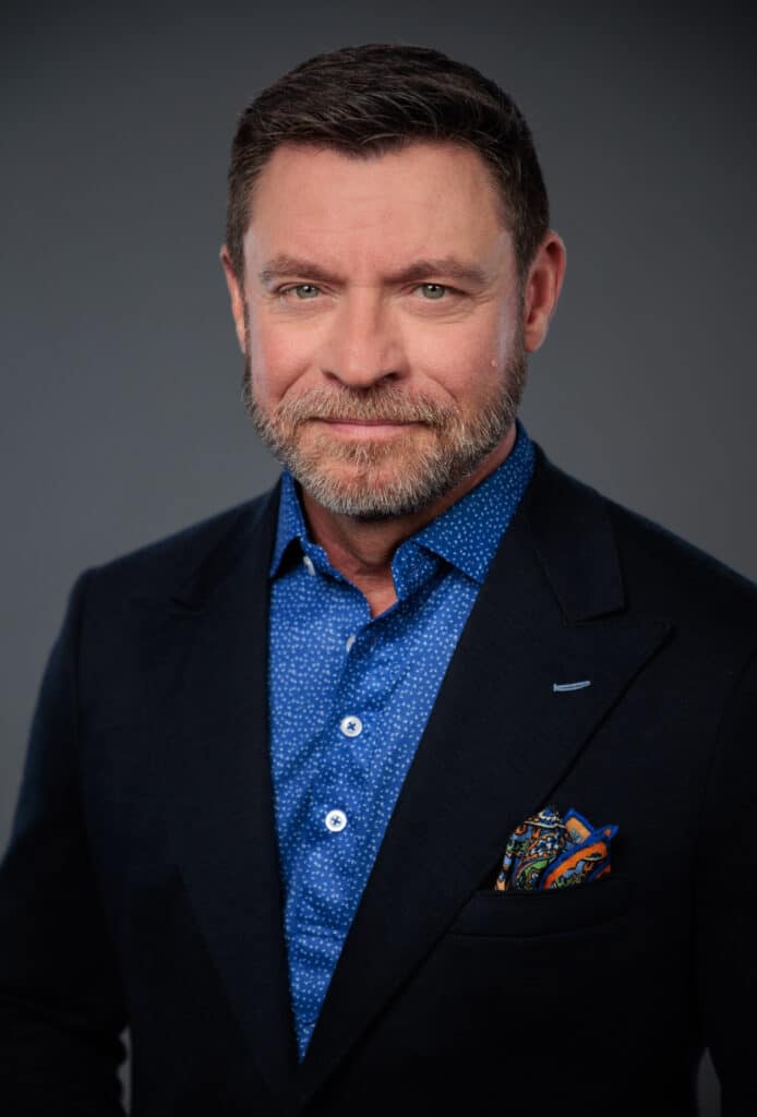 Portrait of Kevin Brown, a middle-aged man with a beard, wearing a suit jacket and a blue patterned shirt, posing against a gray background. He has a serious expression.