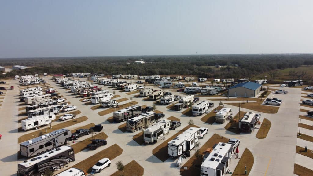 Aerial view of a large RV park with numerous recreational vehicles parked in designated spots, surrounded by sparse vegetation. "Auto Draft