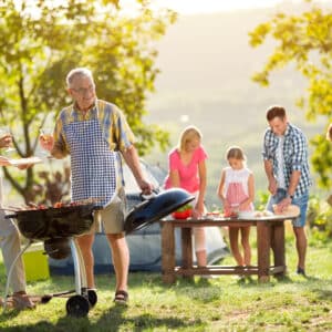A family enjoys a barbecue in a sunny park, with an elderly man grilling, a woman eating, and others preparing food for their exploratory travel.