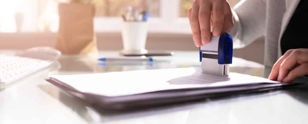 A person stamps a stack of documents on a clear desk. A keyboard, pen, and cup of pens are visible in the background, reminiscent of organizing plans for an exciting trip to Yogi Bear's Jellystone Park with its fun water attractions and glamping options.