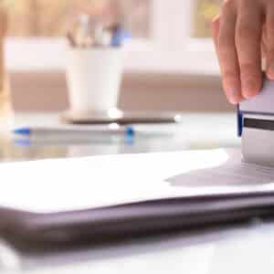 A person stamps a stack of documents on a clear desk. A keyboard, pen, and cup of pens are visible in the background, reminiscent of organizing plans for an exciting trip to Yogi Bear's Jellystone Park with its fun water attractions and glamping options.