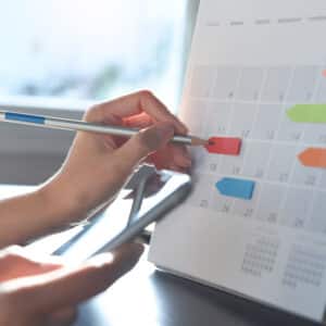 A person using a pen to mark a date on a desk calendar while holding a smartphone in the other hand, planning for festivals and seasonal events. The calendar has colored sticky notes on several dates to attract guests.