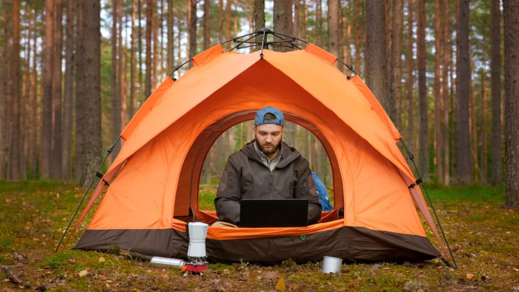 Person sitting inside an orange tent in a forest, working on a laptop. Camping equipment like a portable stove and cup are visible outside the tent. The scene is set among tall trees.