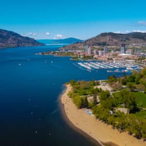 Aerial view of a coastal city with blue water, a sandy beach, greenery, and a marina full of boats. Hills and mountains are visible in the background under a clear blue sky, offering a stunning glimpse of Okanagan's natural beauty—a perfect blend for BC Parks enthusiasts looking to make campsite bookings.