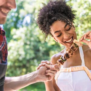 At a vibrant campground, a woman in a white top smiles while enjoying a skewer of grilled meat and vegetables. A man in a plaid shirt and apron holds her hand, both embracing the local culinary experiences outdoors.