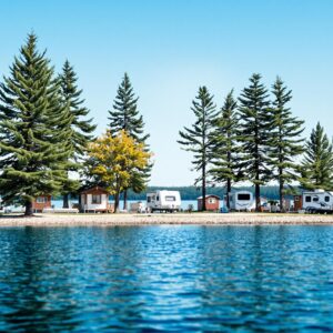 A serene lakeside camping area with RVs and cabins surrounded by evergreen trees under a clear blue sky, featuring a calm shoreline and a relaxed outdoor atmosphere.