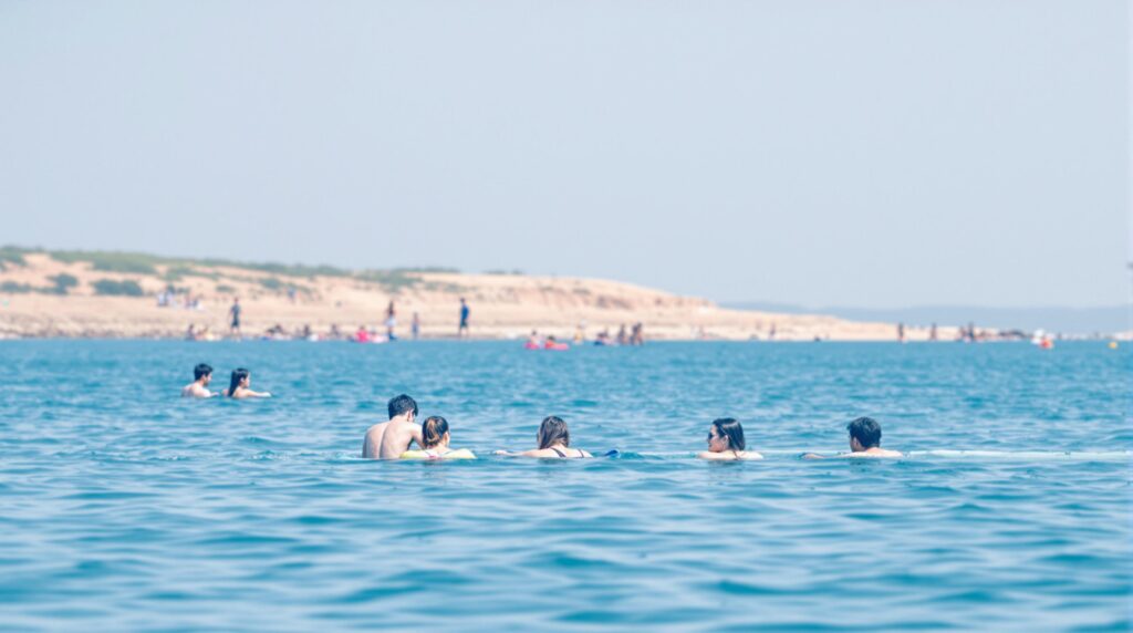 A group of people enjoying recreational activities near a tranquil waterfront, with calm blue water and a sunny, clear sky in the background.
