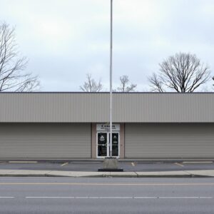 A closed store building with a tall, unmarked flagpole in front under a cloudy sky, surrounded by neutral and nondescript surroundings.
