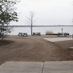 A locked chain barrier blocks access to a deserted lakeside recreation area with empty picnic tables under an overcast sky, surrounded by indistinct foliage in muted colors.