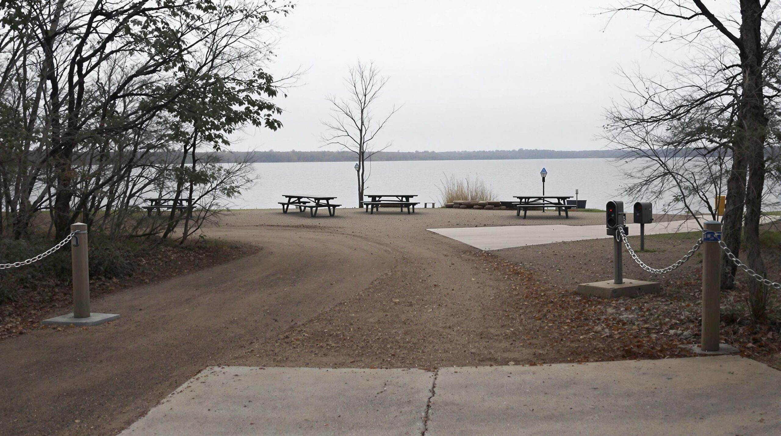 A locked chain barrier blocks access to a deserted lakeside recreation area with empty picnic tables under an overcast sky, surrounded by indistinct foliage in muted colors.