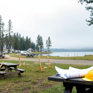 A quiet lakeside campground with survey stakes, a rolled-up blueprint, and a hard hat on a picnic table, RVs and cabins among trees, and a misty lake in the background under an overcast sky.