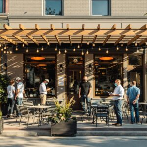 Workers install new patio furniture and pergola outside an anonymous restaurant as the owner reviews plans, with soft sunlight and a blurred urban background.