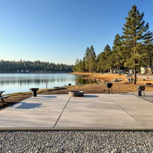 Renovated lakeside campground with new concrete RV pad, picnic table, fire ring, and grill overlooking a calm lake and pine trees, with cabins and subtle construction cones in the background under warm morning light