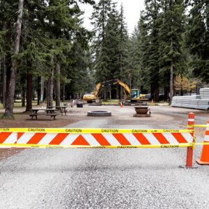 A temporary construction barricade and orange cones block a gravel campground entrance, with caution tape, a picnic table, fire ring, and utility pipes set among evergreen trees. An excavator is visible in the background, indicating infrastructure upgrades.