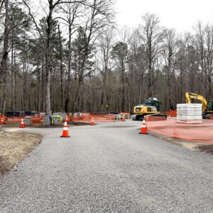Renovation scene at a closed campground loop with empty RV pads, orange construction fencing, a yellow excavator, new picnic tables, and leafless trees in a generic forested area