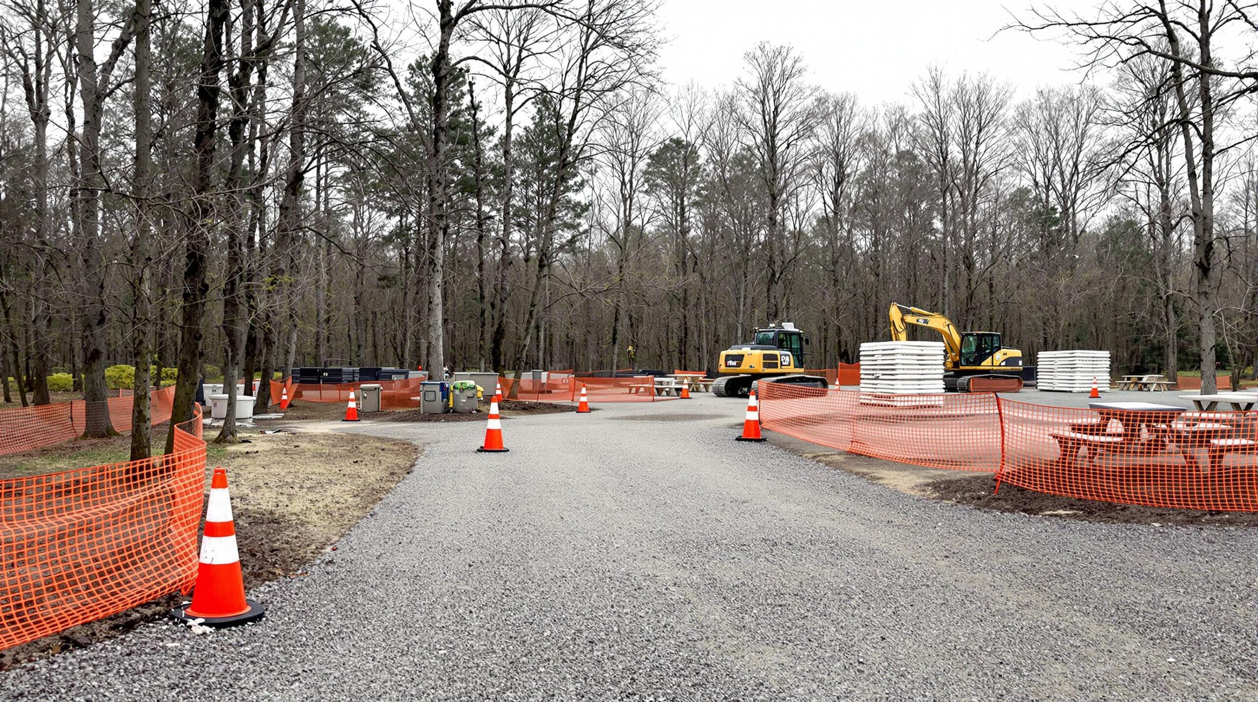 Renovation scene at a closed campground loop with empty RV pads, orange construction fencing, a yellow excavator, new picnic tables, and leafless trees in a generic forested area