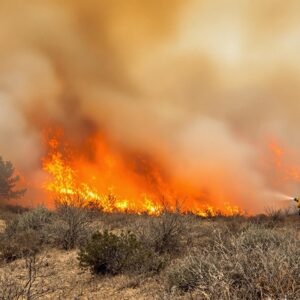 Firefighter in yellow gear battles active wildfire in remote, smoky forested hills with flames and heavy smoke under a hazy sky.