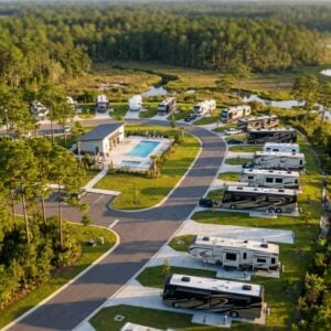 Aerial view of a luxury RV resort featuring paved roads, concrete pads with parked motorhomes and campers, and a central clubhouse with a swimming pool, all surrounded by dense pine forest and a winding marshy river.