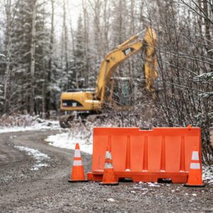 Orange construction barriers and traffic cones block a snowy gravel road next to a wooden sign reading Campground Closed For Renovation, with a yellow excavator visible in the forest background.