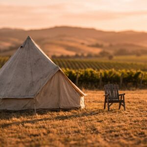 Canvas bell tent and wooden chair situated in a dry field in front of vineyard rows and rolling hills during a warm golden sunset