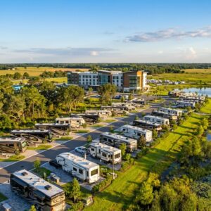 Aerial view of a luxury RV resort and campground featuring paved sites filled with motorhomes and travel trailers next to a scenic wetland lake and boardwalk, with a large modern hotel complex visible in the background under a sunny sky.