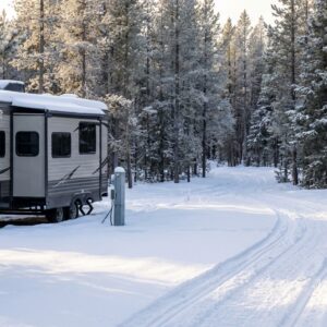 Travel trailer RV parked at a campsite in a snowy pine forest with snow piled on its roof and tire tracks on the adjacent road