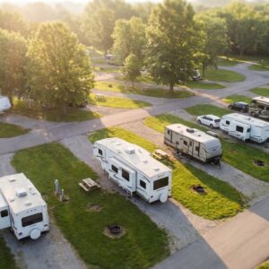 Aerial view of a peaceful RV park campground bathed in warm golden hour sunlight, featuring paved roads winding through gravel campsites occupied by travel trailers and motorhomes surrounded by lush green grass and trees.