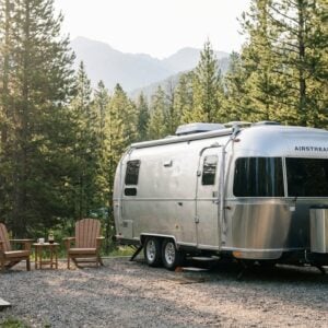 Silver Airstream travel trailer parked on a gravel campsite surrounded by tall pine trees with mountains in the distance, featuring two wooden Adirondack chairs and a side table sitting in the sunlight