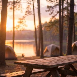 Wooden picnic table in the foreground of a peaceful campground with tents set up among pine trees overlooking a calm lake during a golden sunset