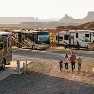 Family of four holding hands walking through a gravel RV park surrounded by luxury motorhomes and travel trailers with scenic red rock mesas in the background during sunset