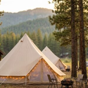 Large canvas glamping bell tents illuminated by string lights sit nestled among tall pine trees next to a rustic log cabin, with camp chairs arranged around a fire pit and forested mountains in the background