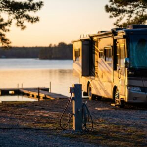 Luxury Class A motorhome parked at a scenic waterfront campsite with full hookups during sunset, featuring a wooden dock extending into the calm lake and pine trees framing the view