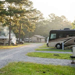 Gravel road running through a wooded RV campground with tall pine trees, featuring a silver Airstream trailer and large brown motorhome parked on the right and campfire smoke rising near campers on the left under morning sunlight.