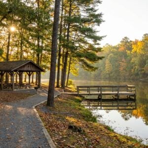 Gravel path leading past a wooden picnic shelter toward a dock on a calm lake surrounded by colorful autumn forest foliage with a sunburst peaking through the trees