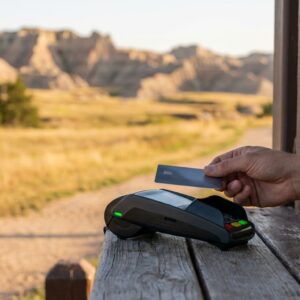Close-up of a hand making a contactless credit card payment on a digital terminal at an outdoor wooden kiosk, with a scenic, out-of-focus landscape of rugged hills and grasslands in the background.