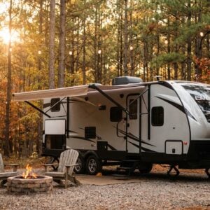 Modern silver travel trailer parked at a gravel campsite in an autumn forest with its awning extended, featuring two wooden Adirondack chairs facing a lit stone fire pit under string lights at sunset.