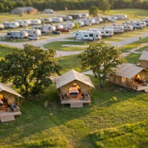 Aerial view of a campground featuring luxury canvas glamping tents on wooden decks in a grassy foreground, with a large RV park full of white campers in the background.