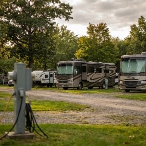 Large Class A motorhomes and travel trailers parked along a gravel road at a wooded RV park, featuring electrical hookup pedestals in the foreground under a cloudy late-afternoon sky