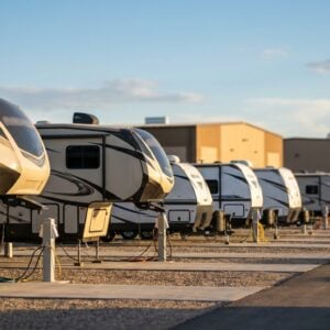 A row of large fifth-wheel recreational vehicles parked on gravel pads and connected to utility hookups at an RV park or dealership lot, with industrial buildings in the background under a partly cloudy blue sky.