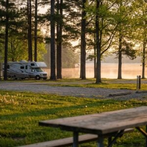 Several RV motorhomes parked at a scenic lakeside campground under tall trees during a misty golden sunrise, with a wooden picnic table on a grassy lawn in the foreground.