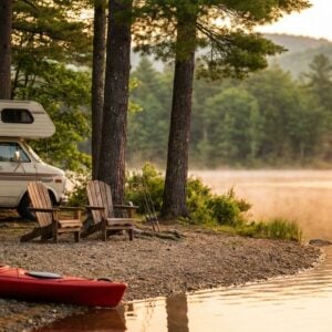 White RV parked at a misty lakeside campsite during a golden sunrise, featuring a red kayak on the rocky shore, two wooden Adirondack chairs facing the calm water, fishing poles leaning against a pine tree, and a picnic table loaded with hiking backpacks.