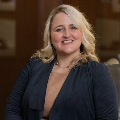 A smiling woman in a black jacket enjoying a cozy fireside chat.