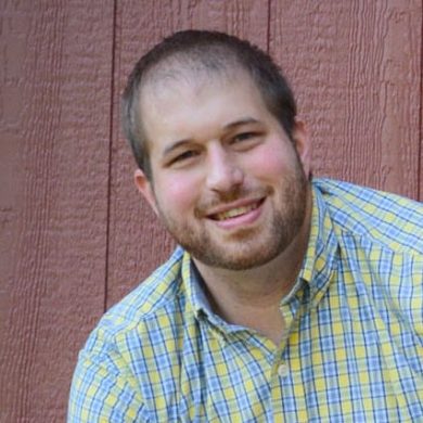 A man in a plaid shirt is smiling in front of a wooden wall during MC Fireside Chats.