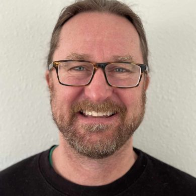 MC Fireside Chats - A man with glasses and a beard smiles in front of a white wall, ready to engage in warm conversations.