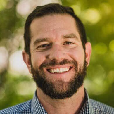 A man with a beard smiling in front of trees, radiating the warm vibes of Fireside Chats.